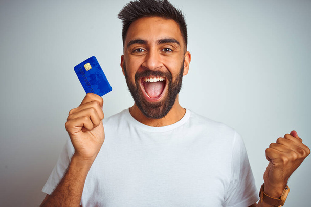 Молодий індійський клієнт тримає кредитну картку, що стоїть над ізольованим білим тлом, кричить гордо і святкує перемогу і успіх дуже схвильовані, веселі емоції
 - Фото, зображення