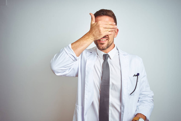 Young handsome doctor man wearing white profressional coat over isolated background smiling and laughing with hand on face covering eyes for surprise. Blind concept. - Photo, Image
