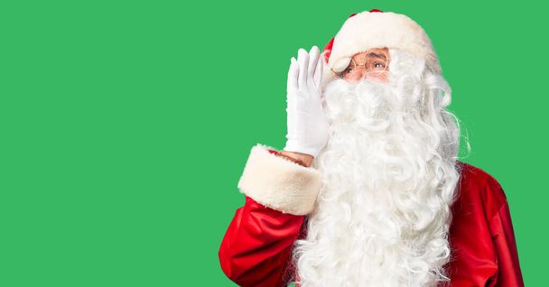 Ein gutaussehender Mann mittleren Alters in Weihnachtsmann-Kostüm und Bart steht schreiend und schreiend mit der Hand auf dem Mund zur Seite. Kommunikationskonzept. - Foto, Bild