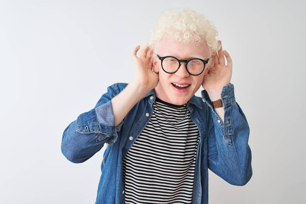 Joven hombre rubio albino con camisa de mezclilla y gafas sobre fondo blanco aislado Tratando de escuchar a ambas manos en el gesto de la oreja, curioso por chismes. Problema de audición, sordos
 - Foto, Imagen