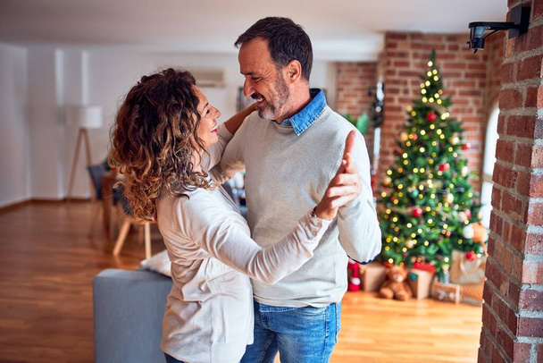 Orta yaşlı güzel çift mutlu ve kendinden emin gülümsüyor. Evde Noel ağacının etrafında dans edip durmak. - Fotoğraf, Görsel