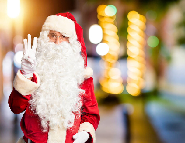 Średniego wieku przystojny mężczyzna w kostiumie Świętego Mikołaja i brodzie stoi pokazując i wskazując palcami numer trzy, uśmiechając się pewny siebie i szczęśliwy. - Zdjęcie, obraz