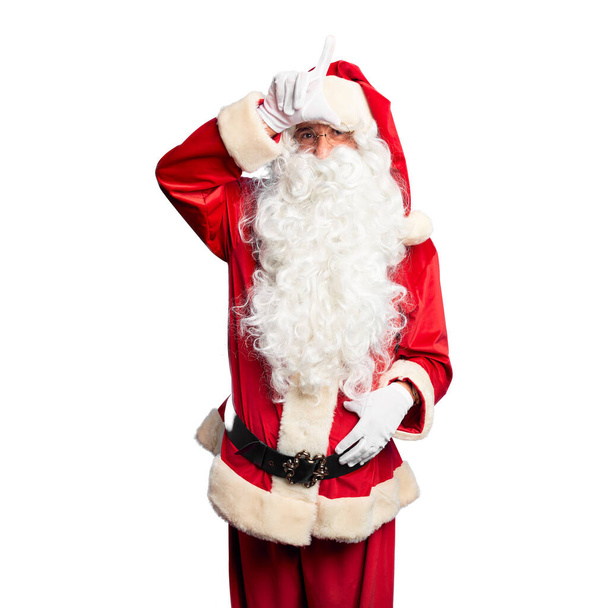 Красивый мужчина средних лет в костюме Санта-Клауса и с бородой стоя высмеивает людей с пальцами на лбу, делая неудачный жест насмешки и оскорбления
. - Фото, изображение