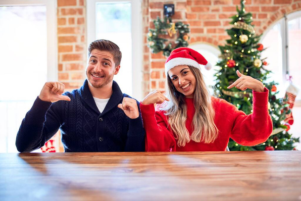 junges Paar mit Weihnachtsmannmütze, das zu Hause auf Stuhl und Tisch um den Weihnachtsbaum sitzt und selbstbewusst lächelnd mit den Fingern stolz und glücklich zeigt. - Foto, Bild