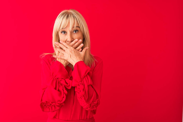 Middle age woman wearing elegant shirt standing over isolated red background shocked covering mouth with hands for mistake. Secret concept. - 写真・画像