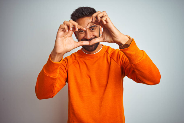 Homem indiano jovem vestindo camisola laranja sobre fundo branco isolado sorrindo no amor fazendo forma de símbolo do coração com as mãos. Conceito romântico
. - Foto, Imagem
