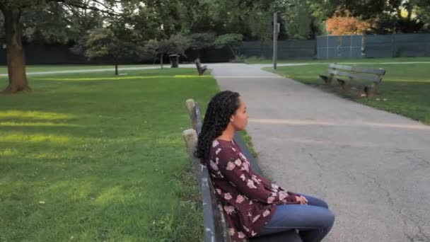Slow motion 180 degree panning walk around a beautiful mixed race African American woman sitting on a weathered black wood and concrete park bench along a path with trees, gardens and grass behind her - Footage, Video