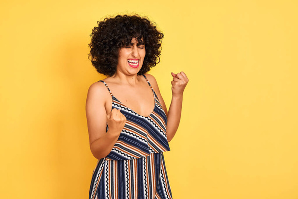 Arab woman with curly hair wearing striped colorful dress over isolated yellow background very happy and excited doing winner gesture with arms raised, smiling and screaming for success. Celebration concept. - Photo, Image
