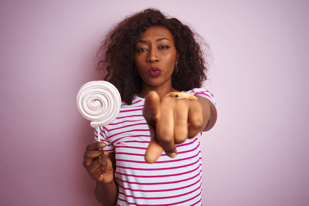 Młoda Afroamerykanka jedząca słodycze stojąca nad odizolowanym różowym tłem wskazującym palcem na kamerę i na ciebie, znak dłoni, pozytywny i pewny siebie gest z przodu - Zdjęcie, obraz