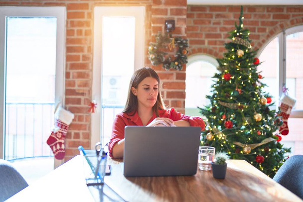 美しい女性座っているテーブルでノートパソコンで作業クリスマスツリーの周りの家手首の時計の時間を確認し、リラックスして自信を持って - 写真・画像