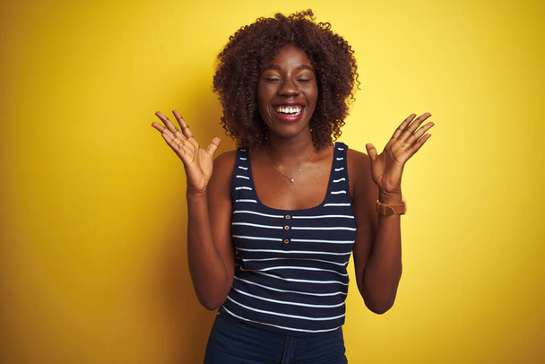 Młoda afrykańska afro kobieta w pasiastej koszulce na odizolowanym żółtym tle świętuje szaleństwo i sukces z podniesionymi ramionami i zamkniętymi oczami krzyczącymi podekscytowani. Koncepcja zwycięzcy - Zdjęcie, obraz