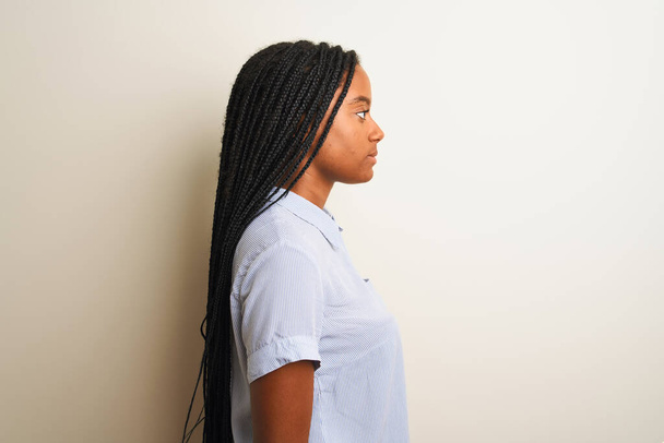 Молодая африканская американка в полосатой рубашке стоя на изолированном белом фоне глядя в сторону, расслабиться профиль позировать с естественным лицом с уверенной улыбкой
. - Фото, изображение