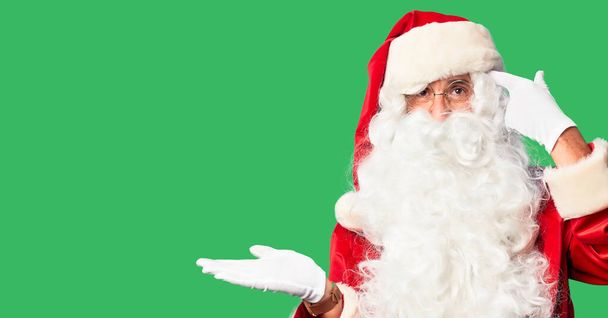 Middelbare leeftijd knappe man in Santa Claus kostuum en baard staan verward en geïrriteerd met open palm tonen kopieerruimte en wijzende vinger naar het voorhoofd. Denk er eens over na.. - Foto, afbeelding