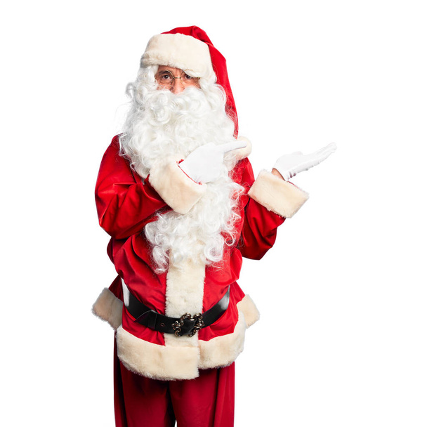 Hombre guapo de mediana edad con traje de Santa Claus y barba de pie asombrado y sonriendo a la cámara mientras se presenta con la mano y señalando con el dedo
. - Foto, imagen
