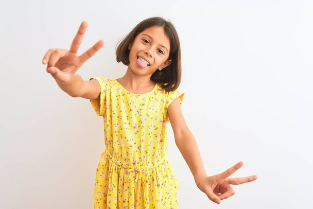 Young beautiful child girl wearing yellow floral dress standing over isolated white background smiling with tongue out showing fingers of both hands doing victory sign. Number two. - Fotó, kép