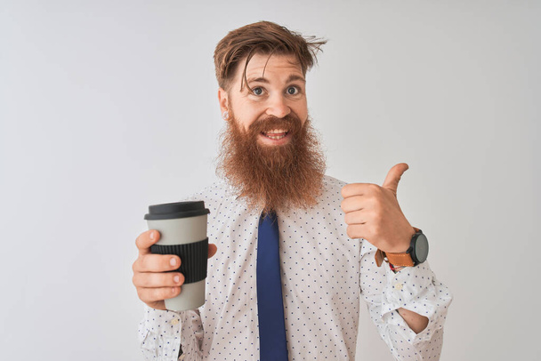 Молодий рудий ірландський чоловік, що п'є, забирає каву, що стоїть над ізольованим білим тлом, вказуючи і показуючи з великим пальцем в сторону з щасливим обличчям посміхаючись
 - Фото, зображення