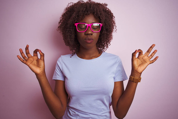 孤立したピンクの背景にTシャツの眼鏡をかけている若いアフリカのアフロ女性は、指で瞑想のジェスチャーをして閉じて目でリラックスして笑顔。ヨガのコンセプト. - 写真・画像