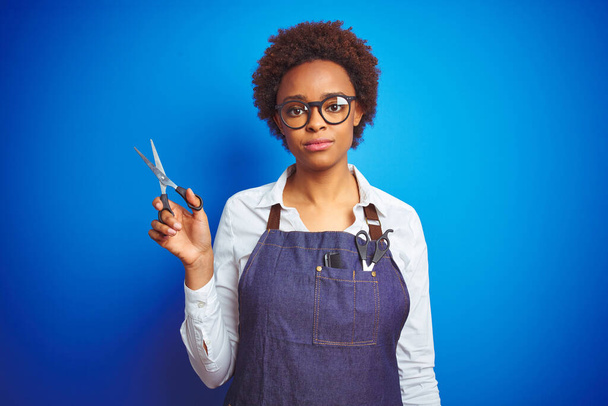 afro-américaine coiffeur femme tenant des ciseaux sur fond bleu isolé avec une expression confiante sur la pensée intelligente visage grave
 - Photo, image