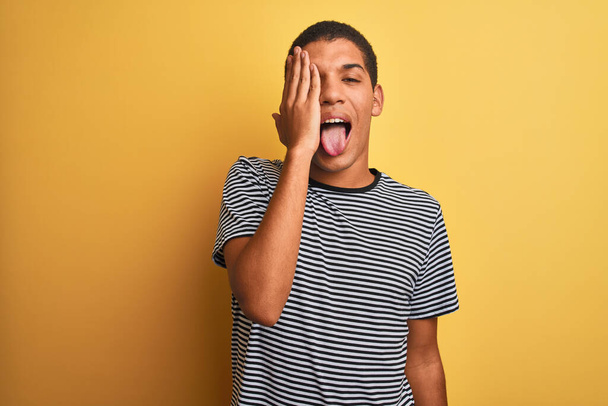 Young handsome arab man wearing navy striped t-shirt over isolated yellow background covering one eye with hand, confident smile on face and surprise emotion. - Photo, Image