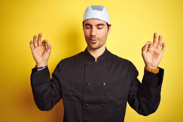 Giovane cuoco bello uomo cucina indossando uniforme e cappello su sfondo giallo isolato rilassarsi e sorridere con gli occhi chiusi facendo gesto di meditazione con le dita. Concetto yoga
. - Foto, immagini