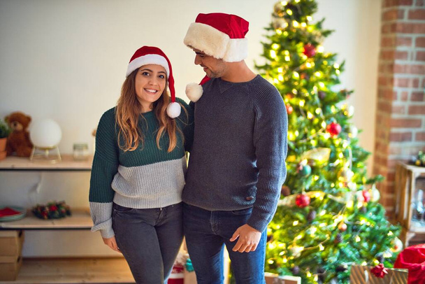 Nuori kaunis pari hymyilee onnellisena ja itsevarmana. Joulupukin hattu päällään seisomassa joulukuusen ympärillä kotona
 - Valokuva, kuva