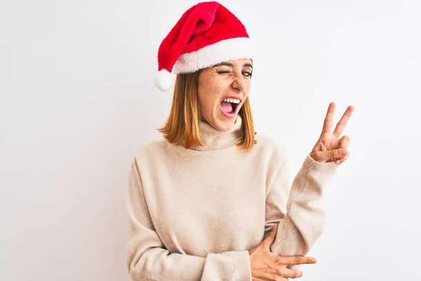 Belle femme rousse portant un chapeau de Noël sur fond isolé souriant avec un clin d'œil heureux à la caméra faisant signe de victoire avec les doigts. Numéro deux
. - Photo, image