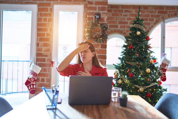 Krásná žena sedící u stolu a pracující s laptopem doma kolem vánočního stromku Dotýkající se čela kvůli nemoci a horečce, chřipce a nachlazení, virové nemoci - Fotografie, Obrázek