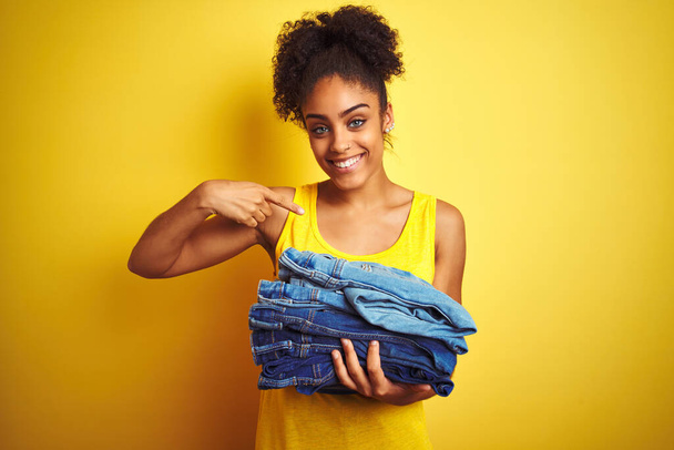 Young african american woman holding stack of jeans over isolated yellow background with surprise face pointing finger to himself - Foto, imagen