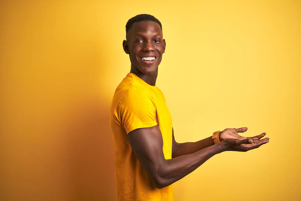 Joven afroamericano hombre vistiendo casual camiseta de pie sobre aislado fondo amarillo señalando a un lado con las manos abiertas palmeras mostrando espacio de copia, presentando anuncio sonriendo emocionado feliz
 - Foto, imagen