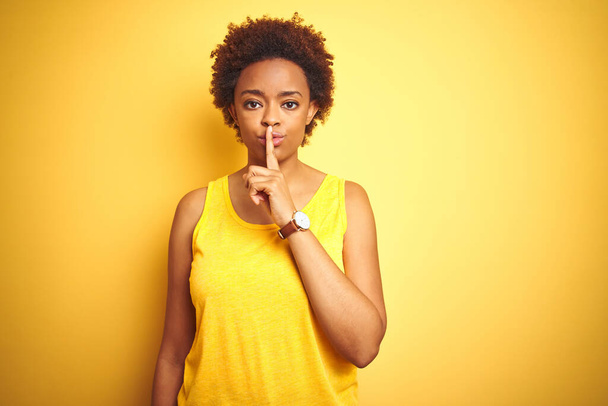 Beauitul donna afro-americana che indossa una t-shirt estiva su uno sfondo giallo isolato chiedendo di essere silenzioso con le dita sulle labbra. Silenzio e concetto segreto
. - Foto, immagini