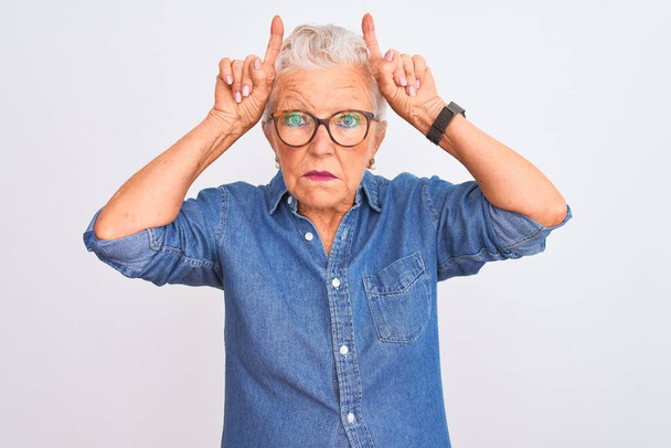 Femme âgée aux cheveux gris portant une chemise en denim et des lunettes sur un fond blanc isolé faisant un geste drôle avec un doigt sur la tête comme des cornes de taureau
 - Photo, image