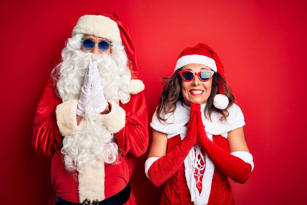 Couple d'âge moyen portant costume de Père Noël et des lunettes de soleil sur fond rouge isolé priant avec les mains ensemble demandant pardon souriant confiant
. - Photo, image