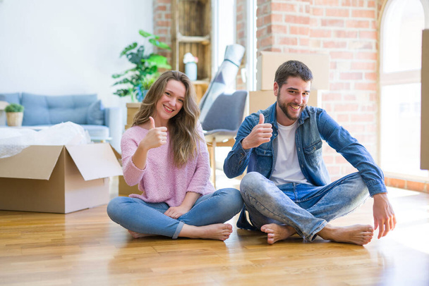 Young beautiful couple moving to a new house sitting on the floor doing happy thumbs up gesture with hand. Approving expression looking at the camera with showing success. - Foto, afbeelding