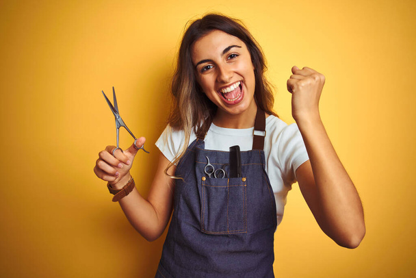 Young beautiful hairdresser woman holding scissors over yellow isolated background screaming proud and celebrating victory and success very excited, cheering emotion - Photo, Image