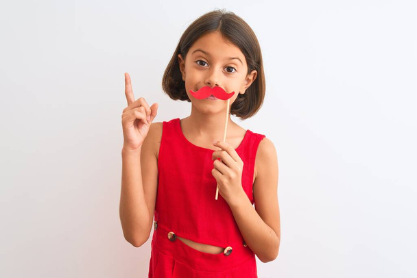 Beautiful child girl holding fanny party mustache standing over isolated white background surprised with an idea or question pointing finger with happy face, number one - Photo, Image