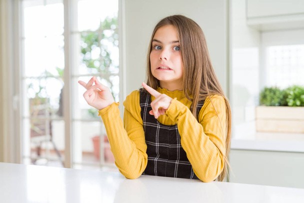 Young beautiful blonde kid girl wearing casual yellow sweater at home Pointing aside worried and nervous with both hands, concerned and surprised expression - 写真・画像