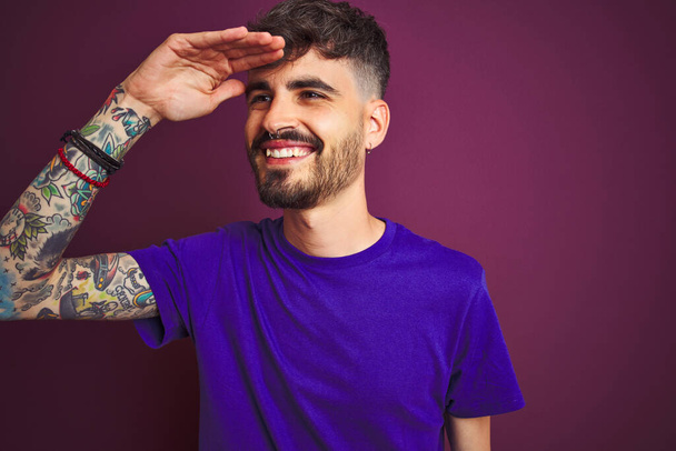 Joven hombre con tatuaje vistiendo camiseta de pie sobre fondo púrpura aislado muy feliz y sonriente mirando lejos con la mano sobre la cabeza. concepto de búsqueda
. - Foto, imagen