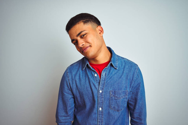 Young brazilian man wearing denim shirt standing over isolated white background looking away to side with smile on face, natural expression. Laughing confident. - Foto, immagini