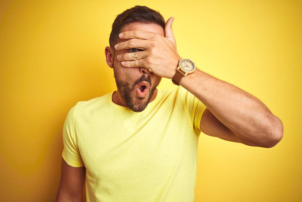 Young handsome man wearing casual yellow t-shirt over yellow isolated background peeking in shock covering face and eyes with hand, looking through fingers with embarrassed expression. - 写真・画像