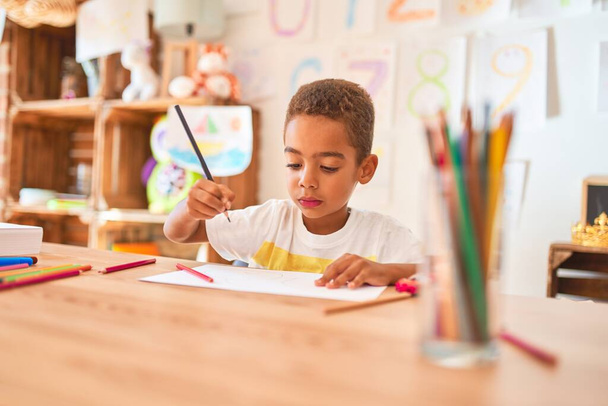Beautiful african american toddler sitting drawing using paper and pencils on desk at kindergarten - Фото, изображение