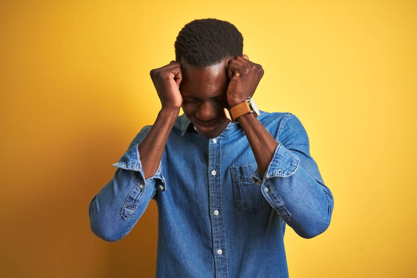 Молодий афроамериканець, одягнений в джинсову сорочку, стоїть над ізольованим жовтим фоном, який страждає від головного болю і стресу через біль і мігрень. Руки за голову. - Фото, зображення
