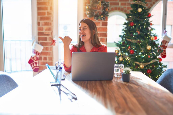 Mooie vrouw zitten aan de tafel werken met laptop thuis rond kerstboom glimlachen met gelukkig gezicht kijken en wijzen naar de zijkant met duim omhoog. - Foto, afbeelding