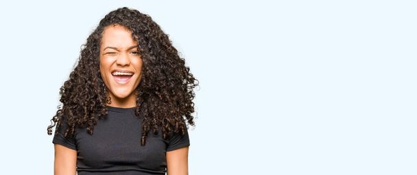 Young beautiful woman with curly hair winking looking at the camera with sexy expression, cheerful and happy face. - Photo, Image