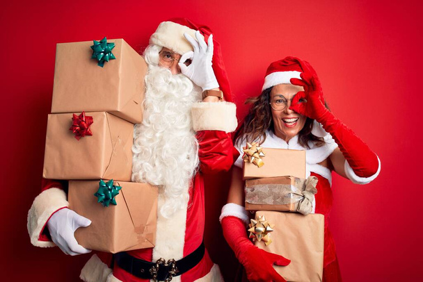 Middle age couple wearing Santa costume holding tower of gifts over isolated red background doing ok gesture with hand smiling, eye looking through fingers with happy face. - Photo, Image