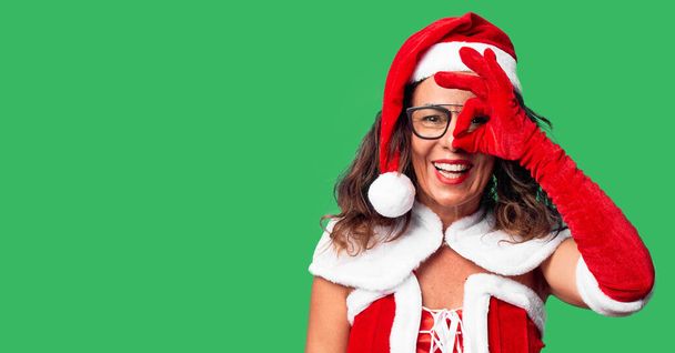 Μεσήλικη γυναίκα φορώντας κοστούμι Άγιος Βασίλης κάνει εντάξει χειρονομία με το χέρι χαμογελώντας, τα μάτια κοιτάζοντας μέσα από τα δάχτυλα με χαρούμενο πρόσωπο. - Φωτογραφία, εικόνα