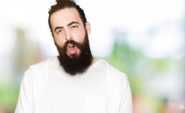 Young hipster man with long hair and beard wearing sporty sweatshirt In shock face, looking skeptical and sarcastic, surprised with open mouth - Foto, afbeelding