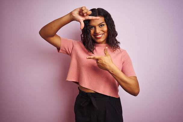 Jonge mooie transseksuele transgender elegante vrouw over geïsoleerde roze achtergrond glimlachen maken frame met handen en vingers met gelukkig gezicht. Creativiteit en fotografie concept. - Foto, afbeelding