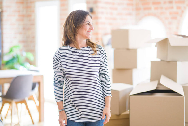 Middle age woman moving to a new house arround cardboard boxes looking away to side with smile on face, natural expression. Laughing confident. - Photo, Image