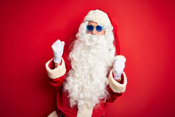 Muž středního věku v kostýmu Santa Clause a slunečních brýlích na izolovaném červeném pozadí velmi šťastný a vzrušený dělá vítězný gesto se zdviženýma rukama, usmívá se a křičí po úspěchu. Koncept oslavy. - Fotografie, Obrázek