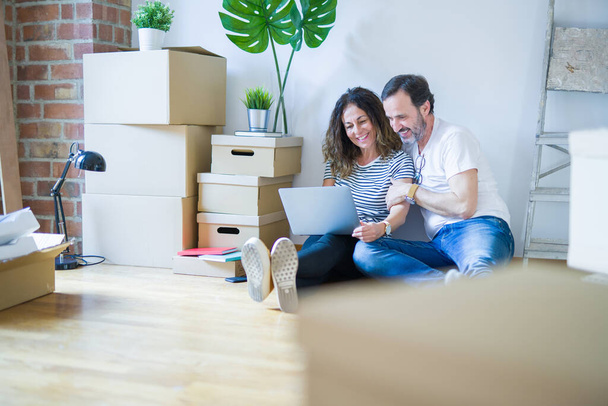 Romantisches Paar mittleren Alters, verliebt, auf dem Boden der Wohnung sitzend, mit Boxen herum und Computer-Laptop lächelnd glücklich für den Umzug in ein neues Zuhause - Foto, Bild